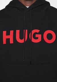 Felpa nera con grande logo rosso "HUGO" attraversato sul petto e lacci neri che pendono dal cappuccio.