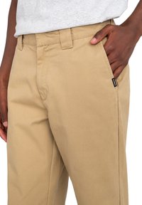 Hnedé chinos nohavice vyrobené z odolnej bavlnenej zmesi. Obsahujú plochý predný diel, bočné vrecká a malé čierne logo značky na vrecku.