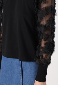 Schwarzes Oberteil mit durchsichtigen, gerafften schwarzen Netzärmeln; fester Körper; taillierte Bündchen; getragen mit einem blauen Denim-Rock; strukturierter Stoff schafft Kontrast.