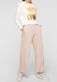 Haut blanc à manches longues avec imprimé graphique doré, associé à un pantalon large en velours côtelé rose pâle et des baskets crème clair.