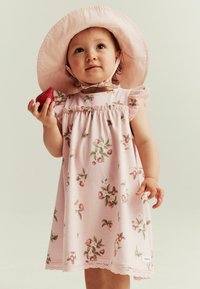 Vaaleanpunainen kukallinen mekko, jossa on lyhyet röyhelöhiha, kerätty pääntiE, pitsireunus ja yhteensopiva vaaleanpunainen aurinkohattu. Lapsi pitää mansikkaa.