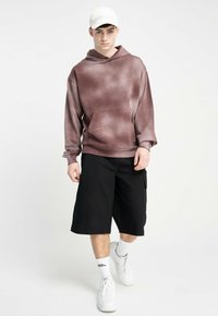 Brązowy hoodie w tie-dye z przednią kieszenią, zestawiony z czarnymi szortami do kolan. Model nosi białą czapkę i białe trampki. Casualowy, luźny krój.