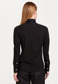 Femme aux cheveux lisses châtain clair portant une chemise noire ajustée à manches longues et un pantalon noir à fines rayures, vue de dos sur un fond blanc.