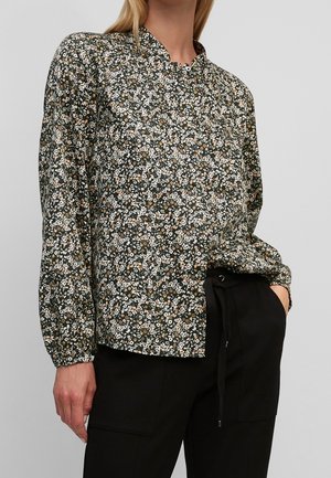 Vrouw draagt een zwarte blouse met lange mouwen met een kleine witte en beige bloemenprint, gecombineerd met zwarte broek met aantrekkoord in de taille.