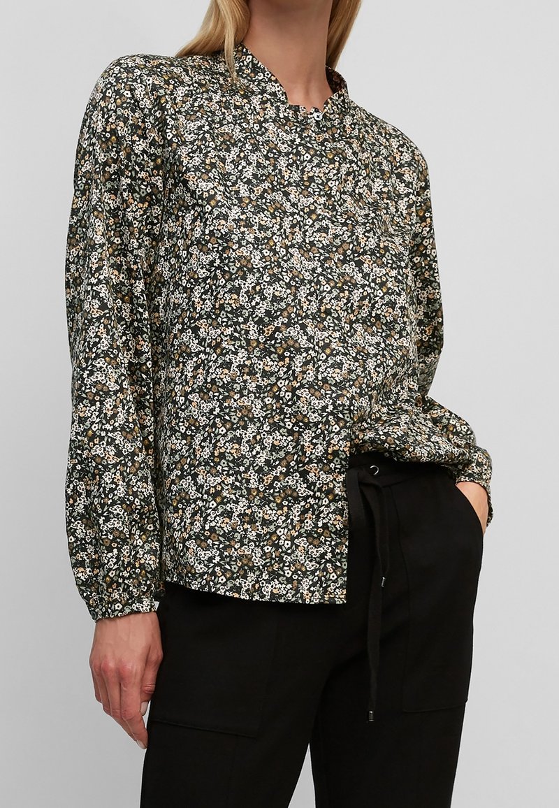 Femme portant un chemisier noir à manches longues avec un petit imprimé floral blanc et beige, associé à un pantalon noir à taille élastiquée avec cordon.