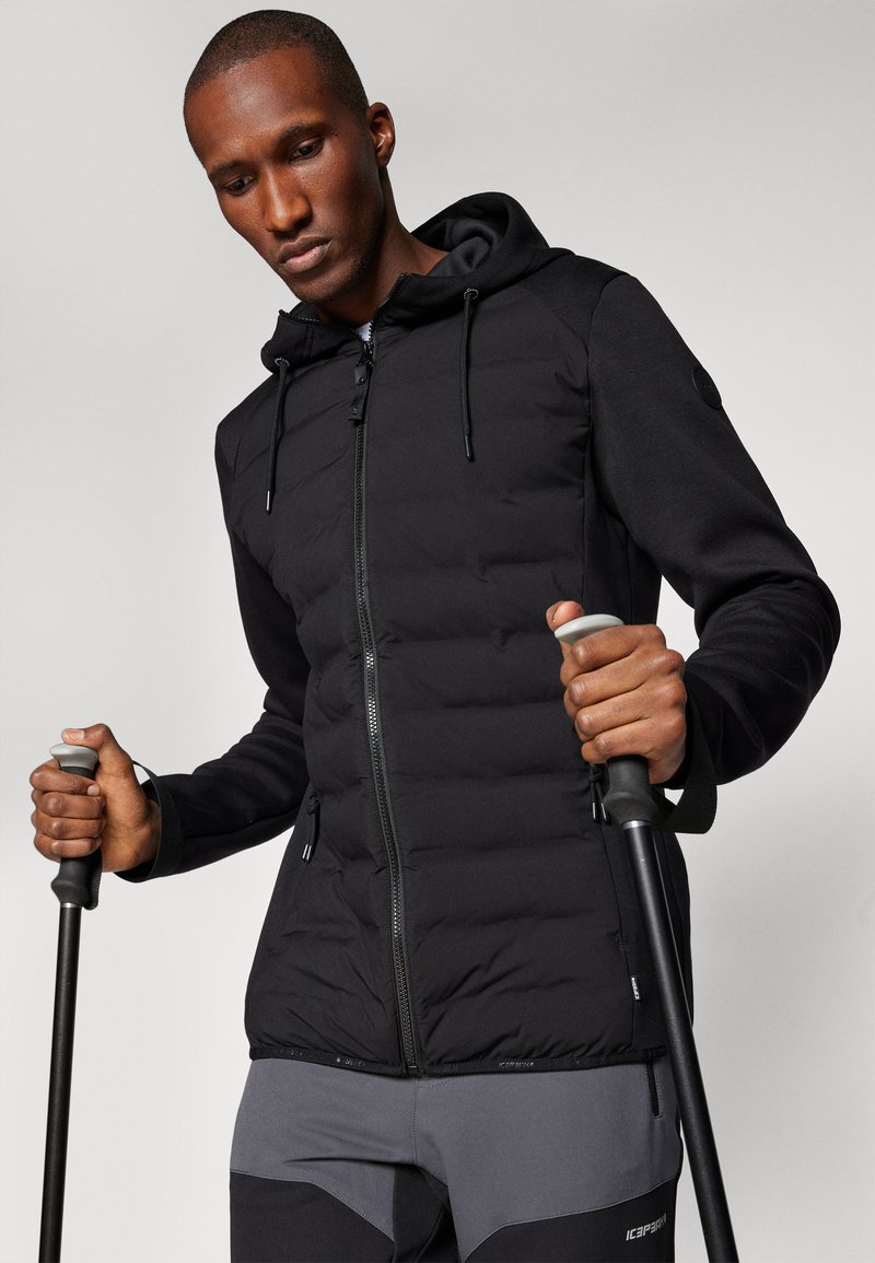 Zwarte gewatteerde jas met een ritssluiting aan de voorkant en een capuchon, met een gladde textuur. Gecombineerd met wandelstokken, gedragen over grijze sportieve broek.