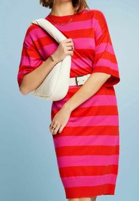 Femme portant une robe rayée rouge et rose avec ceinture blanche, tenant un sac à épaule blanc matelassé sur un fond bleu clair.
