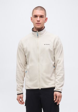 Jeune homme aux cheveux courts portant une veste polaire Columbia beige clair zippée, debout devant un fond blanc uni.