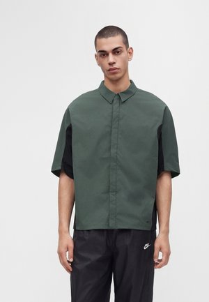 Männliches Model mit kurzen Haaren, das ein lockeres dunkelgrünes kurzärmeliges Hemd mit Knöpfen und schwarze Nike-Hosen vor einem einfarbigen Hintergrund trägt.