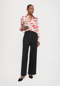 Patterned shirt in pink and red shades with lip motifs, paired with wide black pinstripe trousers and holding a metallic clutch.