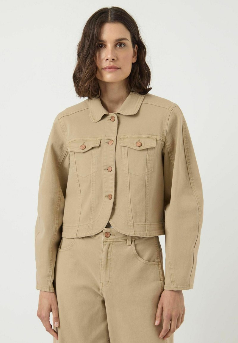 Vrouw met schouderlang bruin haar, die een beige cropped denim jacket draagt met knoopzakken op de borst en bijpassende hoge taillebroek.