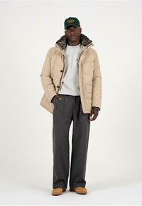 Giacca in nylon color beige con colletto in pelliccia sintetica, bottoni neri, maglione grigio, pantaloni plissettati grigi e scarpe beige; modella in piedi su uno sfondo bianco.