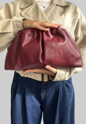 Borsa a pochette in pelle cremisi con design arricciato, texture morbida e finitura liscia, tenuta con entrambe le mani su uno sfondo grigio.