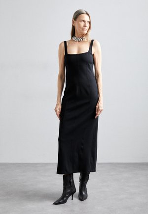 Black fitted dress with square neckline and thin straps. Smooth texture, knee-length. Paired with black ankle-high pointed boots and layered chain necklace.