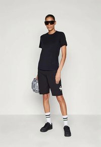 Fekete pólót, fekete kerékpáros sortot, fekete sportcipőt és csíkozott fehér zoknit viselő nő, aki egy szürke kerékpáros sisakot tart, és nagy fekete napszemüveget visel.