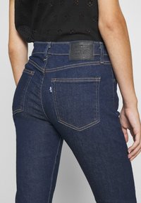Mörkblå denimjeans har en fitted design med synliga sömmar och två bakfickor. Bälteområdet visar en texturerad svart läderlapp.