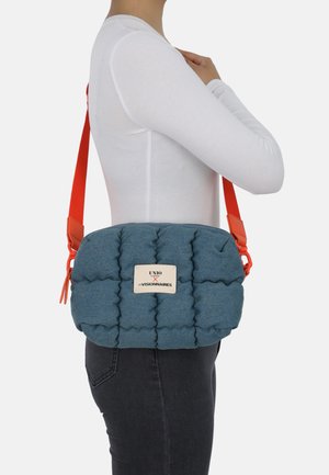 Blaue gepolsterte Umhängetasche mit orangefarbenem Riemen und einer Plakettenbeschriftung. Texturiertes Gewebe mit gesteppter Oberfläche und verstellbarem Riemen.