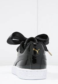 Černé patentní kožené tenisky s bílou podrážkou, které mají velkou černou saténovou mašli, zlaté logo Puma na patě a strukturovanou gumovou podrážku.