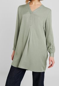Hellgrün, langärmlige Tunika mit V-Ausschnitt und Knopfleiste vorne. Glattes Material, leicht lockere Passform und Raffung an den Seiten.