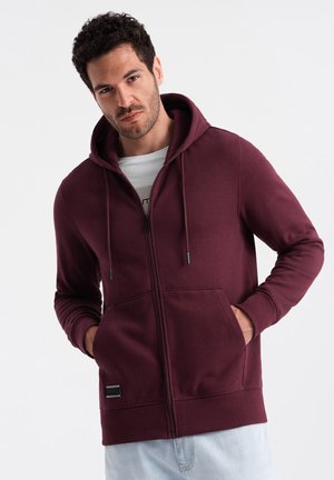 Sudadera con cremallera en burdeos de tejido suave, que cuenta con un bolsillo frontal, capucha con cordón y puños acanalados. Incluye una etiqueta con el logo en el dobladillo.