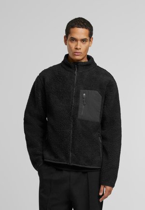 Black fleece jacket with a zip front, high collar, and a chest pocket. Features textured fabric, zippered side pockets, and contrasting accents.