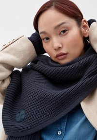 Écharpe tricotée bleu marine avec un motif texturé, superposée à une pièce d'extérieur beige clair et une chemise en denim bleu. Dispose d'un logo noir proéminent.