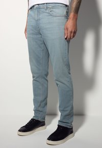 Jean en denim bleu clair à coupe slim, présentant un léger délavage, des poches standard et une fermeture à bouton à la taille, associé à des baskets noires.