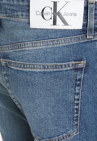 Džíny Calvin Klein, modrá denimová tkanina, rovný střih, detail zadní kapsy, bílá štítek s černým textem, viditelné švy.