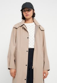 Trench-coat imperméable beige avec boutons-pression et col large, associé à un pull en maille blanc et une casquette de baseball noire.