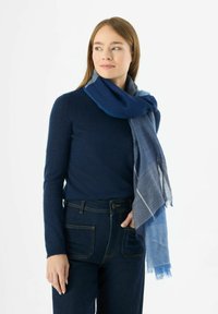 Foulard bleu aux nuances variées, avec une texture douce et des bords effilochés, porté sur un pull sombre. Jean en denim avec des coutures visibles.