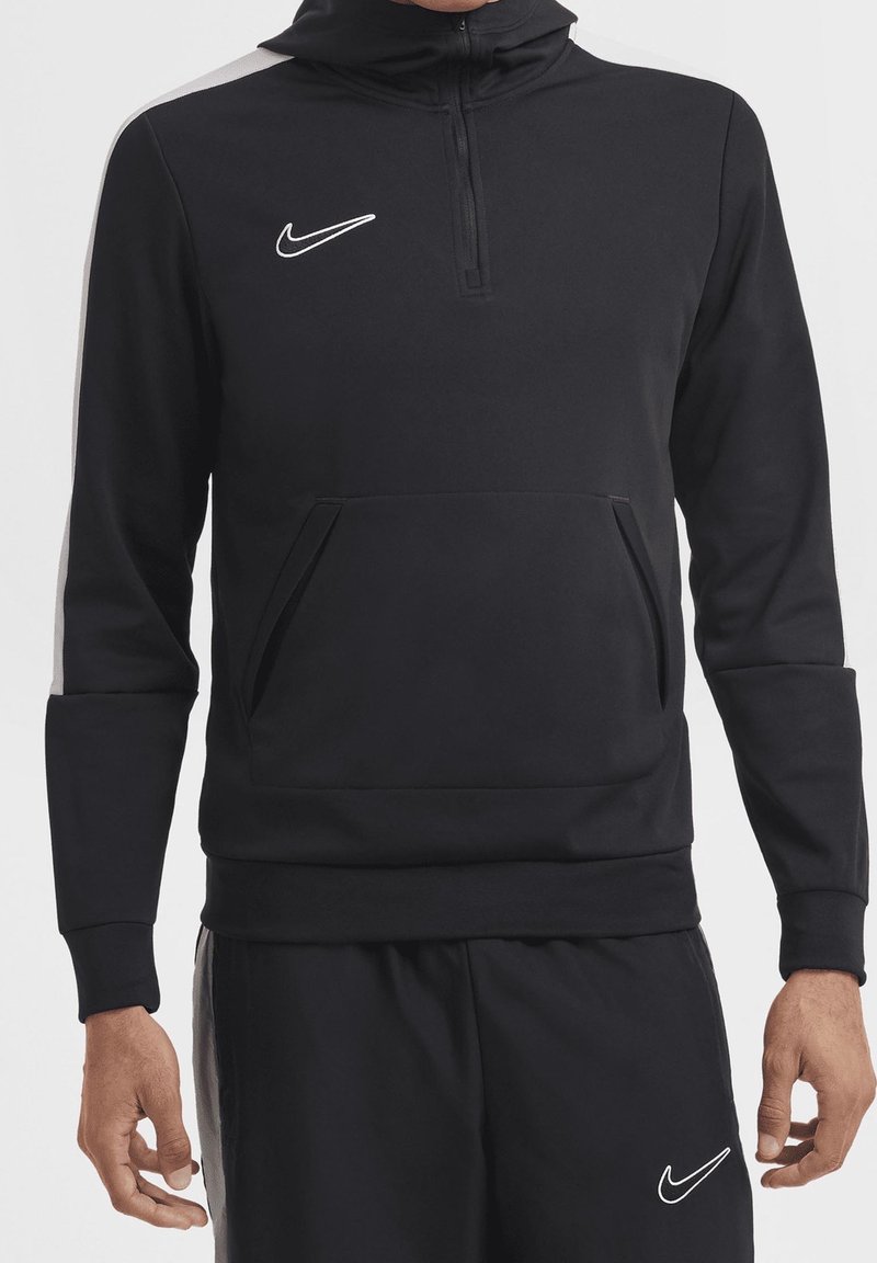 Personne portant un survêtement noir Nike avec des logos swoosh blancs, un sweat à capuche à fermeture éclair mi-longue, et un pantalon assorti avec des poches latérales.