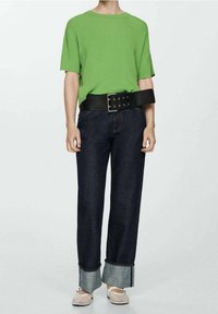 Grünes geripptes Strickshirt mit kurzen Ärmeln, schwarzer breiter Gürtel, dunkle Jeans mit Aufschlägen und hellen Schuhen. Einfaches und strukturiertes Design.