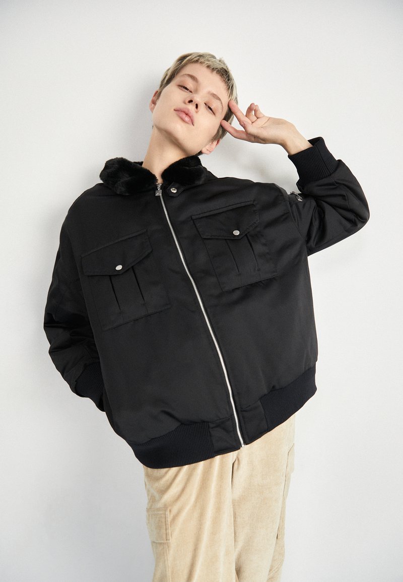 Sort bomberjakke med lynlås foran, imiteret pelskrave og to brystlommer. Har ribbede manchetter og kant samt en glat satinoverflade.