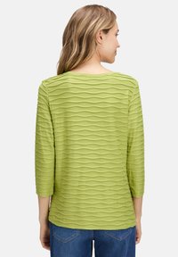 Grüner, texturierter Oberteil mit wellenförmigem Muster, 3/4-Ärmeln und rundem Ausschnitt, kombiniert mit Denim-Jeans. Rückansicht gezeigt.