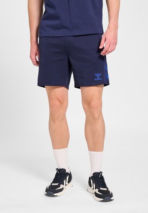 Mand iført marineblå Hummel-shorts, hvide crew-sokker og sorte og hvide atletiksko, stående på en hvid baggrund.