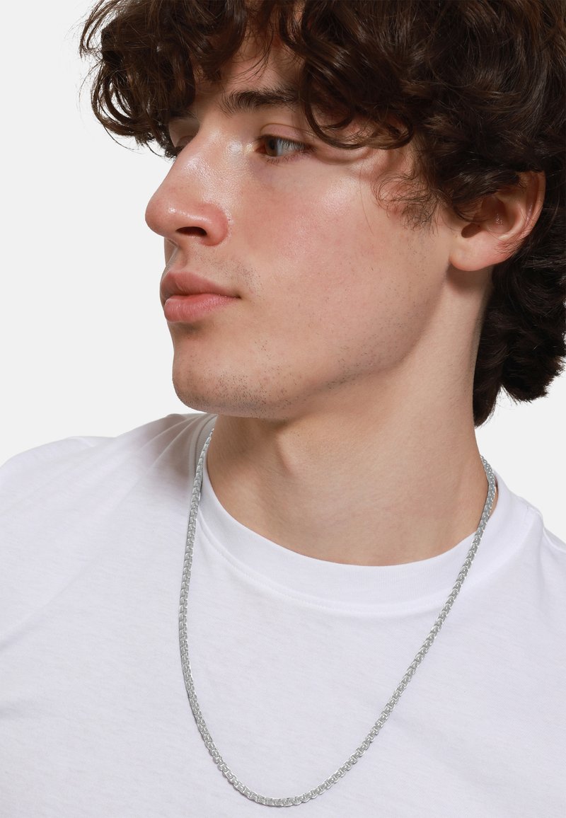 Collana in argento indossata sopra una maglietta bianca semplice, con un design a maglie rotonde. Il metallo ha una finitura lucida e una texture liscia.