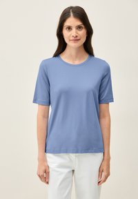 Světle modré tričko s krátkým rukávem a kulatým výstřihem, hladké na omak, v kombinaci s bílými kalhotami. Jednoduchý a minimalistický design.