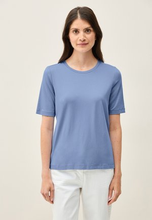 Světle modré tričko s krátkým rukávem a kulatým výstřihem, hladké na omak, v kombinaci s bílými kalhotami. Jednoduchý a minimalistický design.