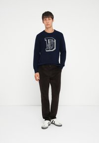 Pull en mailles bleu marine avec une lettre "D" en relief et un motif d'ancre, associé à un pantalon marron foncé et des baskets blanches avec des accents noirs.