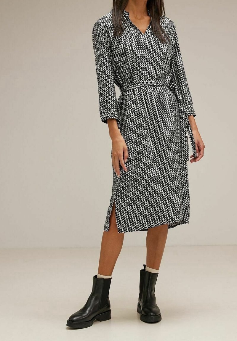 Femme portant une robe midi à motifs noir et blanc avec une ceinture et des bottines noires, debout devant un fond uni.
