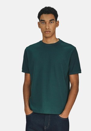 Grünes, strukturiertes T-Shirt mit quadratischem Muster, kurzen Ärmeln und rundem Ausschnitt. Das Model steht mit den Händen in den Taschen. Dunkle Jeans.