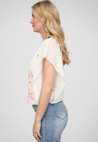 Top bianco leggero e trasparente con maniche a sbuffo e motivo floreale rosa, abbinato a jeans blue attillati, visto di lato.