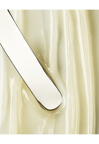 Spatule en métal avec une surface lisse et réfléchissante, partiellement immergée dans une substance crémeuse de couleur jaune pâle avec des textures tourbillonnantes subtiles.