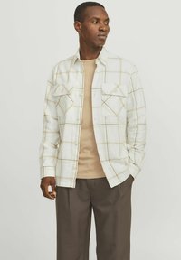 Camisa de cuadros blanca con líneas diagonales, dos bolsillos frontales, diseño de botones. Lleva una parte superior beige debajo y está combinada con pantalones marrones oscuros.
