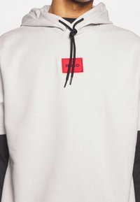 Sudadera con capucha gris con un parche cuadrado de logotipo rojo. Cuenta con cordones negros y detalles en las mangas negros para contraste.