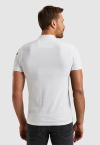 Bijela polo majica kratkih rukava sa mekim teksturom, s ravnim krojem i malim detaljem u obliku aplikacije na stražnjem dijelu vrata, uparena s tamnim trapericama.