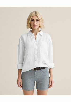 Blond kvinde iført en hvid skjorte med knapper og opkrogede ærmer samt stribede shorts til midt på låret med sort bælte, stående mod en ensfarvet baggrund.