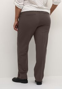 Braune Hose mit glatter Textur, geradem Schnitt und Gesäßtaschen. Kombiniert mit einem hellen, gemusterten Shirt und schwarzen Loafers.