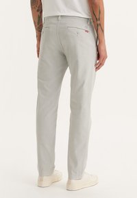 Pantalones gris claro de algodón con corte slim y una textura sutil. Presenta bolsillos traseros y una pequeña etiqueta de logo roja.