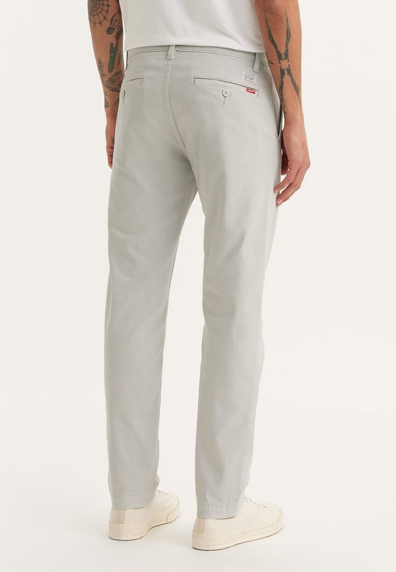 Pantalones gris claro de algodón con corte slim y una textura sutil. Presenta bolsillos traseros y una pequeña etiqueta de logo roja.